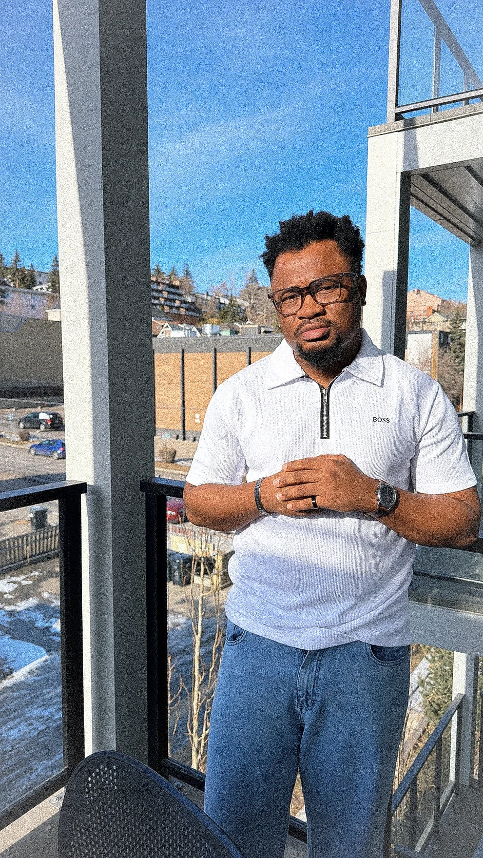 Oluwayemi leaning on a balcony rail in a white zip polo and jeans, on a bright winter day