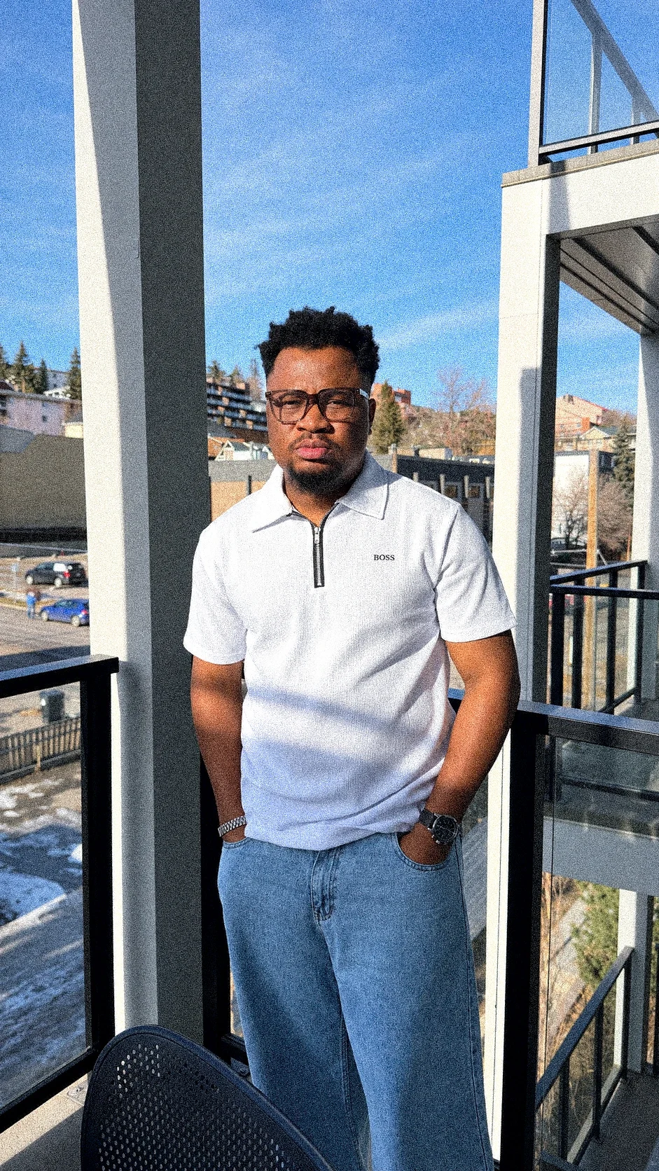 Oluwayemi standing on a balcony with his hands in his pockets, city rooftops and blue sky behind him