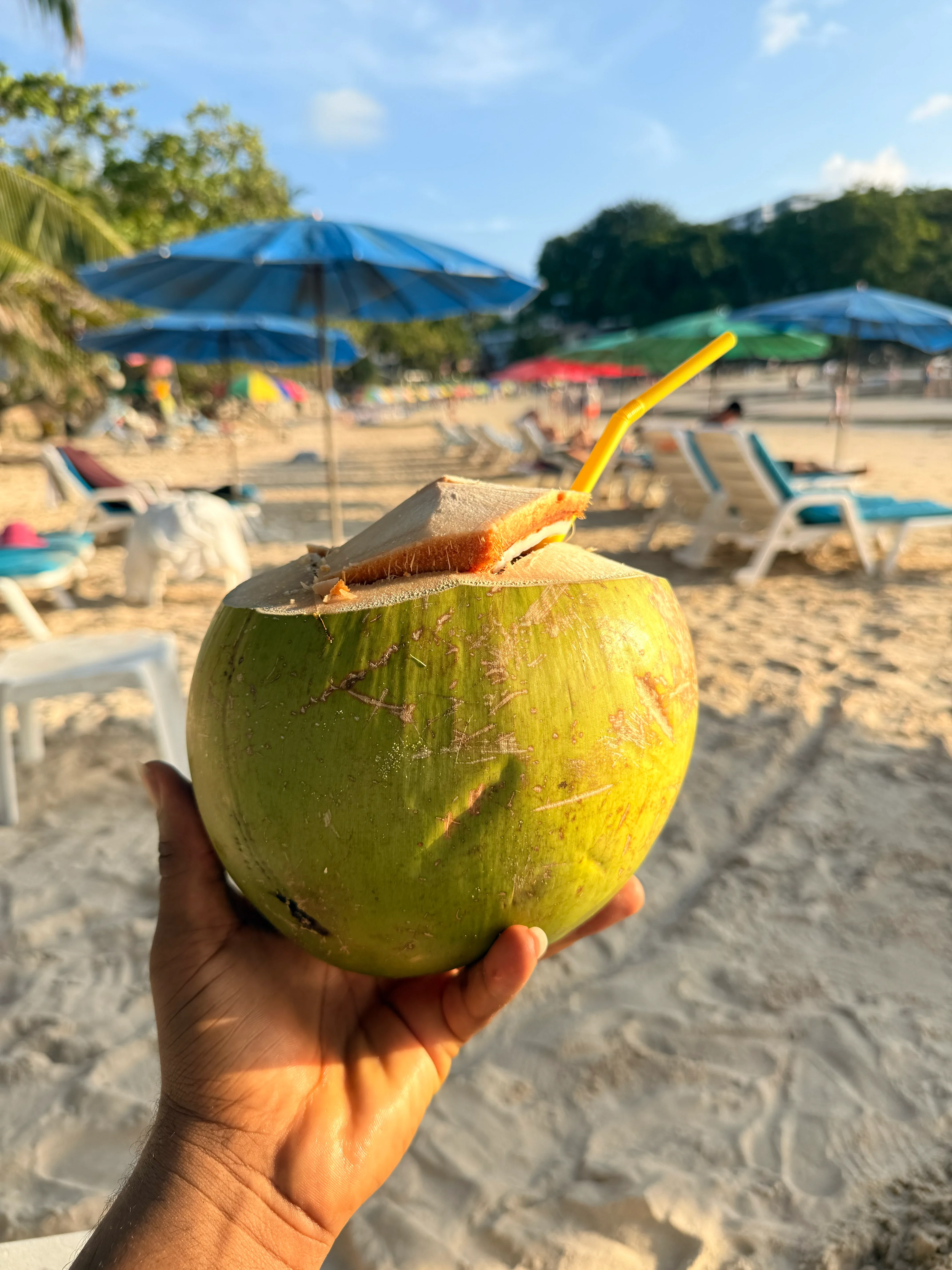 A hand holding up a fresh coconut with a straw, on a sunny beach lined with blue umbrellas and loungers