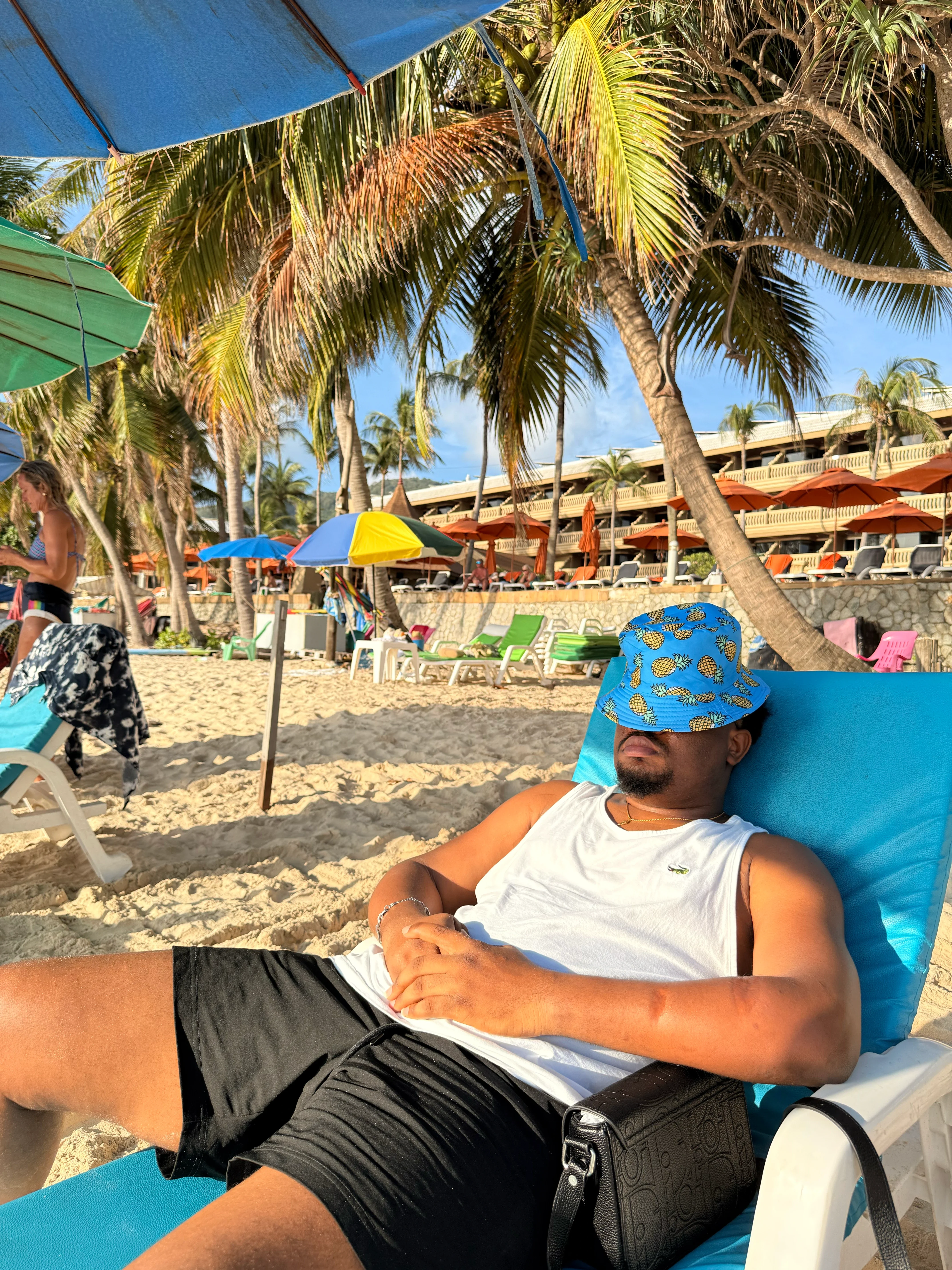 Oluwayemi relaxing on a beach lounger under palm trees, in a bucket hat pulled over his eyes