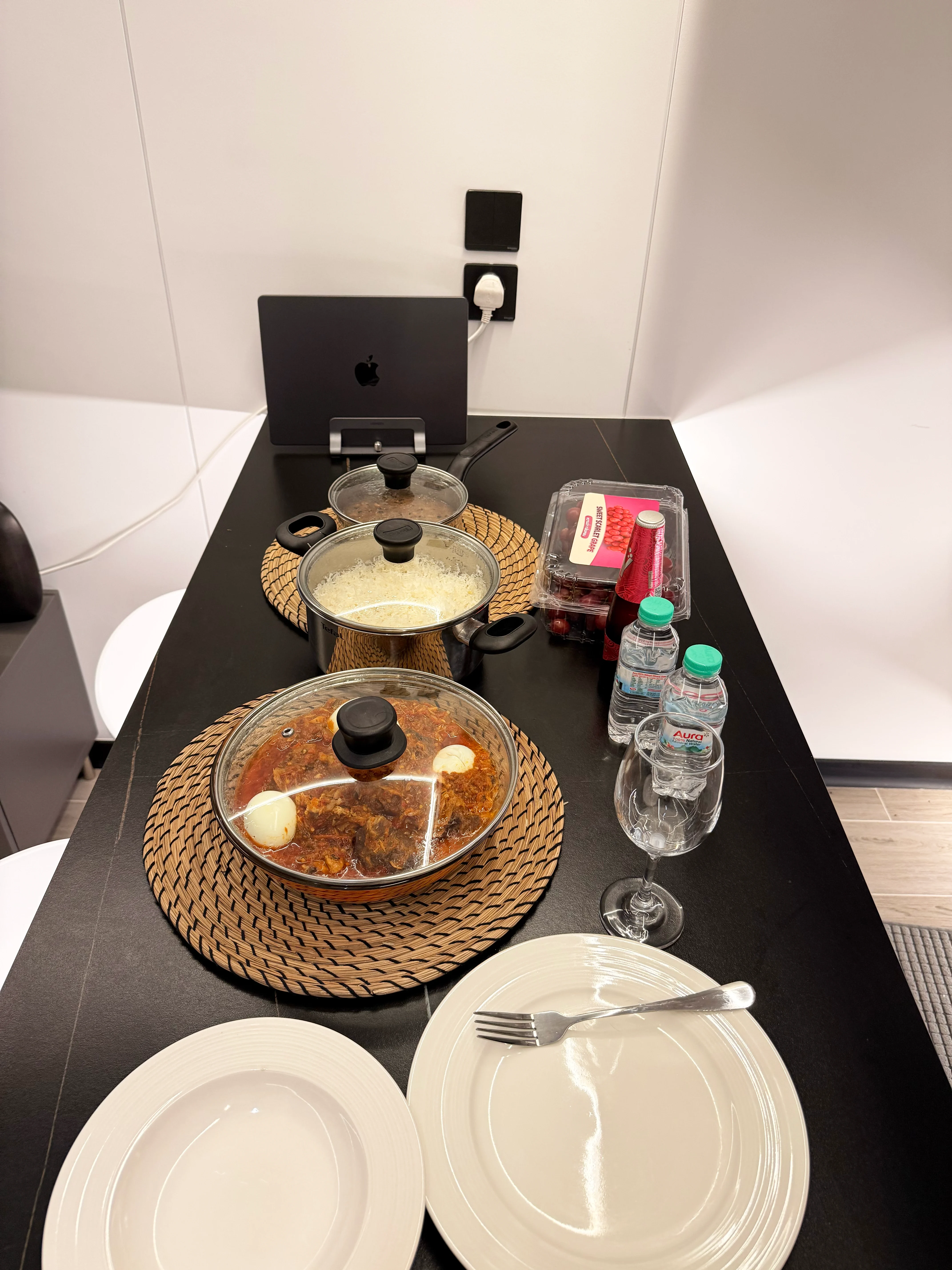 Dinner on a kitchen counter: a pot of rice, a stew in a woven trivet, plates and glasses, with a laptop at the back