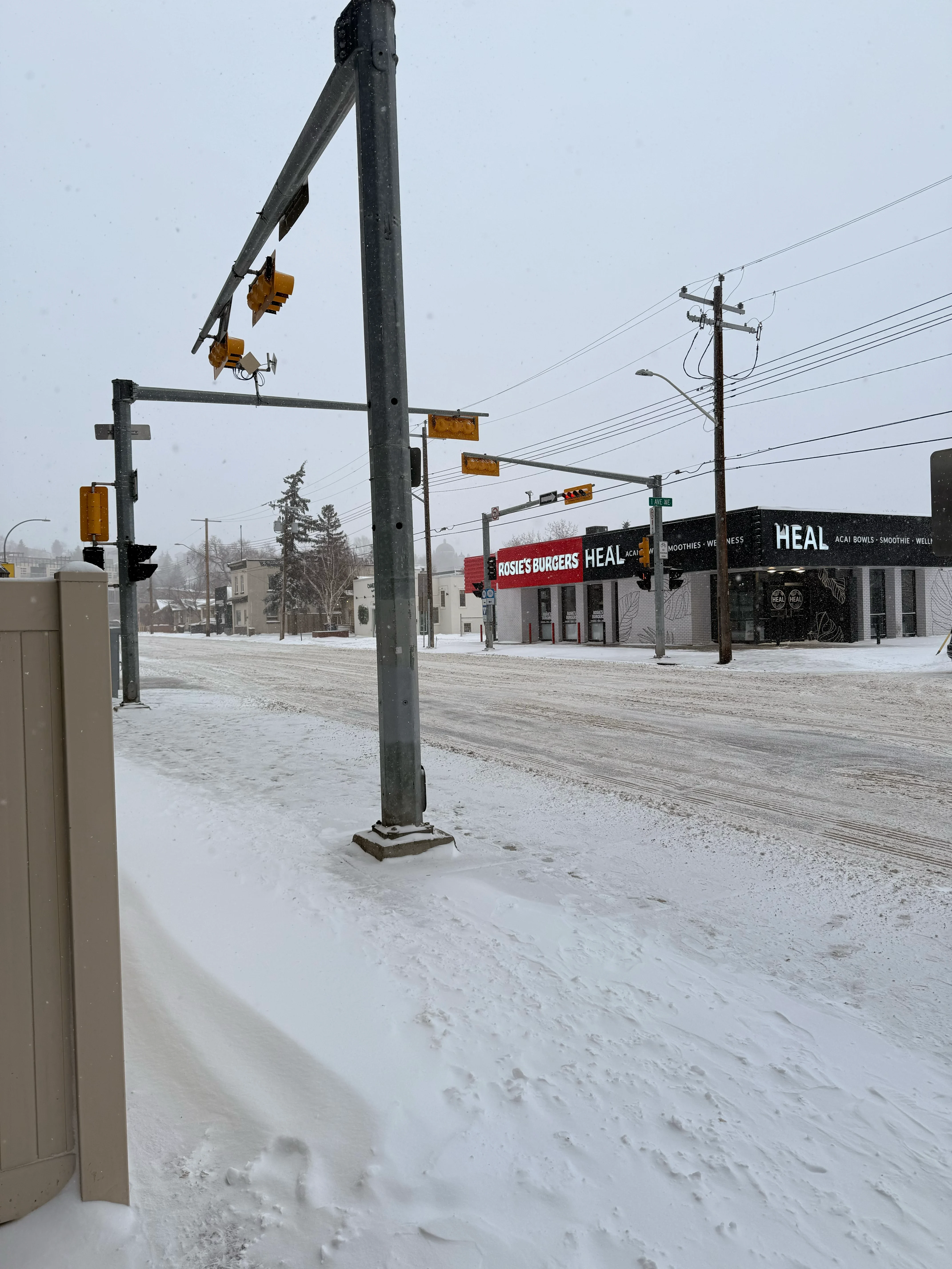 A snowy intersection, traffic lights under falling snow, with a burger shop and a wellness bar on the far corner