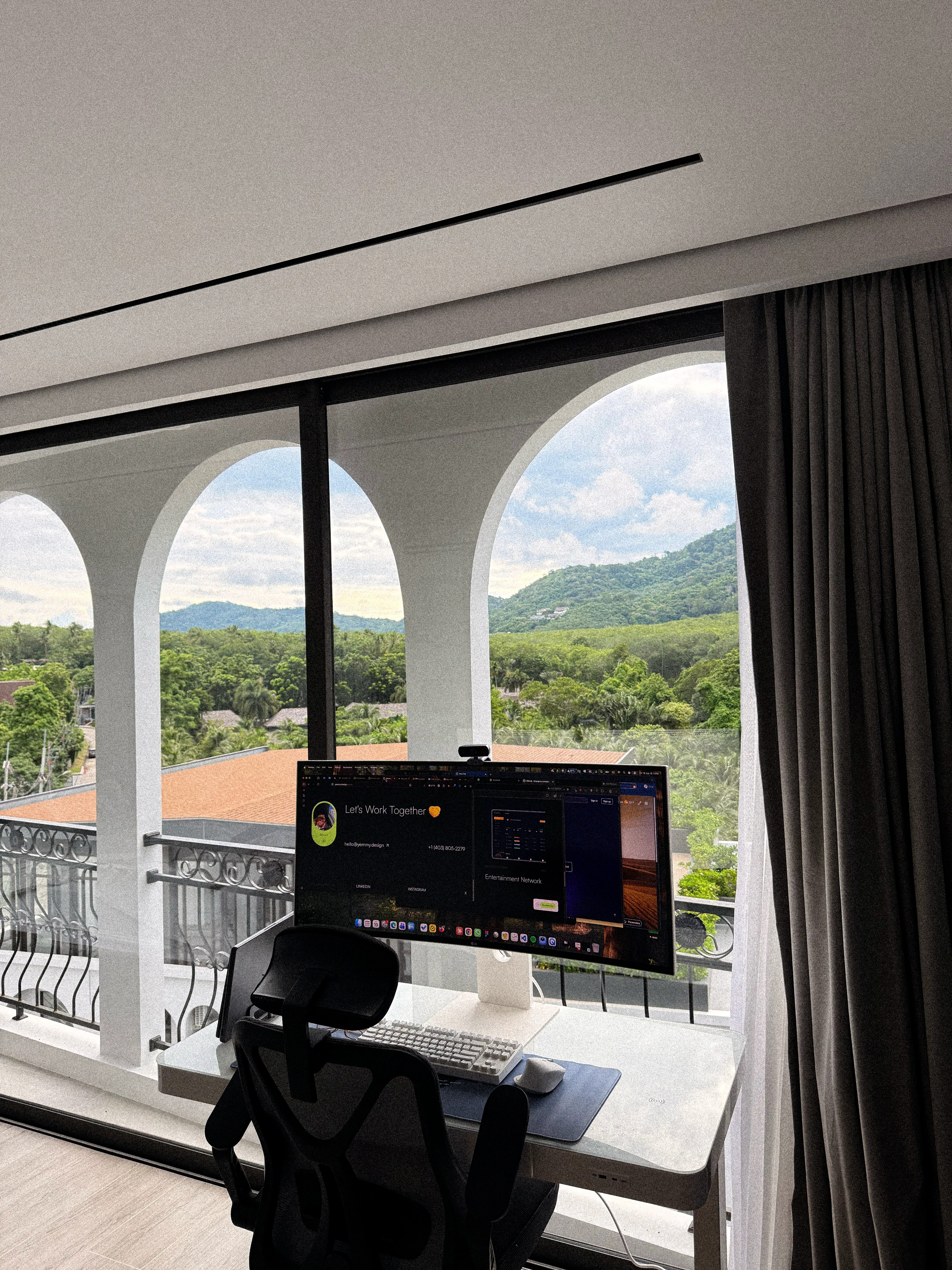 A desk and monitor facing a wide arched window, with green hills outside