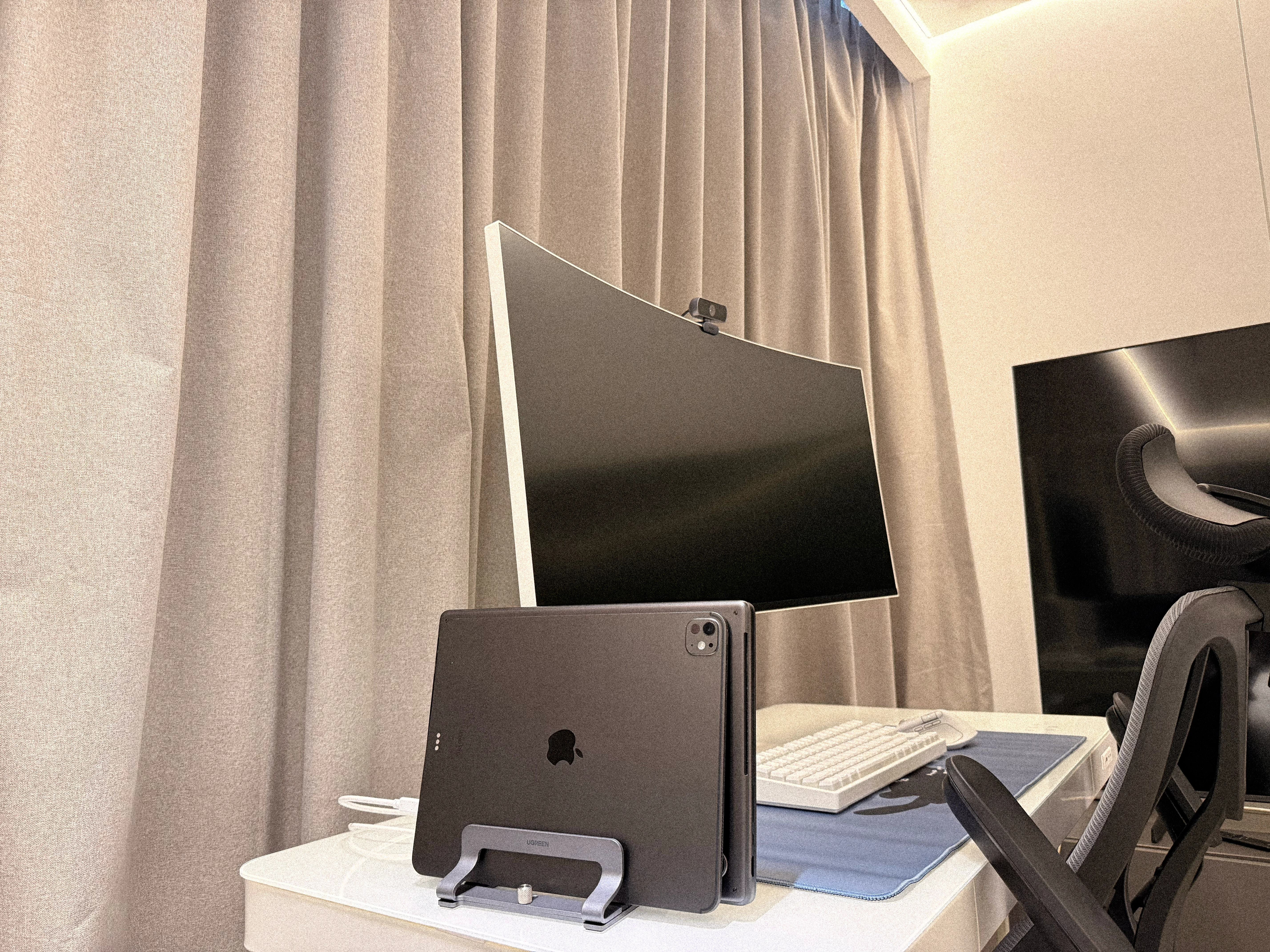 His workstation: a curved monitor, a laptop in a vertical stand and a desk chair beside the curtains