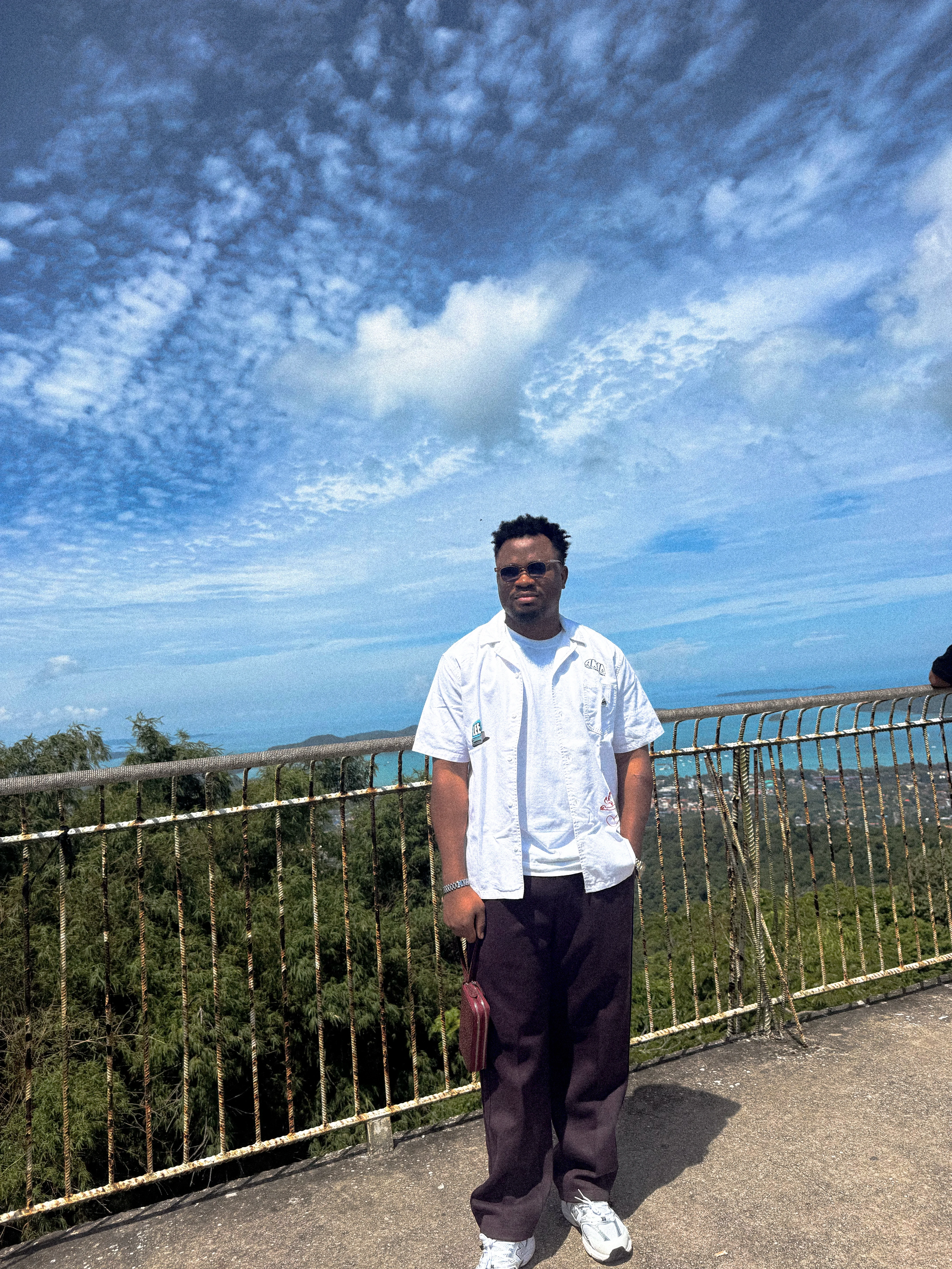 Oluwayemi at a hilltop railing in a white shirt, with the coastline and a wide cloudy sky behind him
