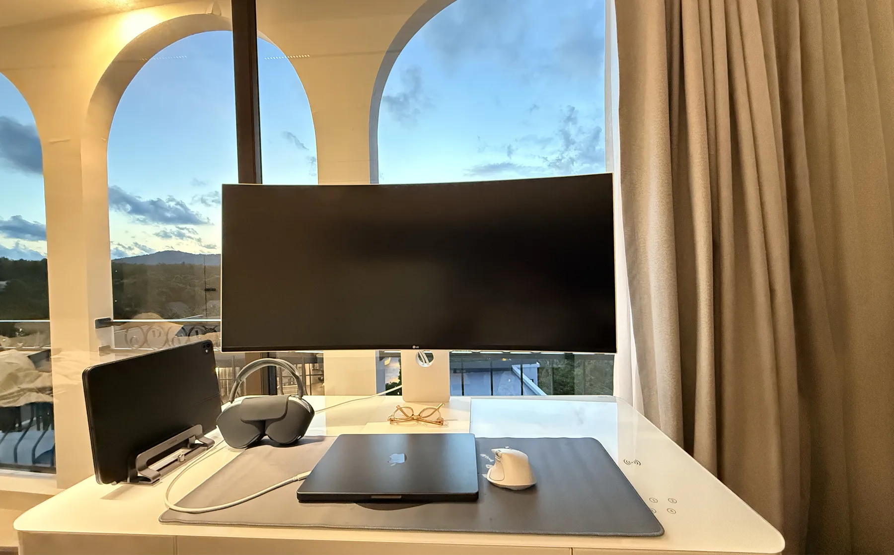 My workstation: a curved monitor, a laptop and headphones at a desk by arched windows at dusk
