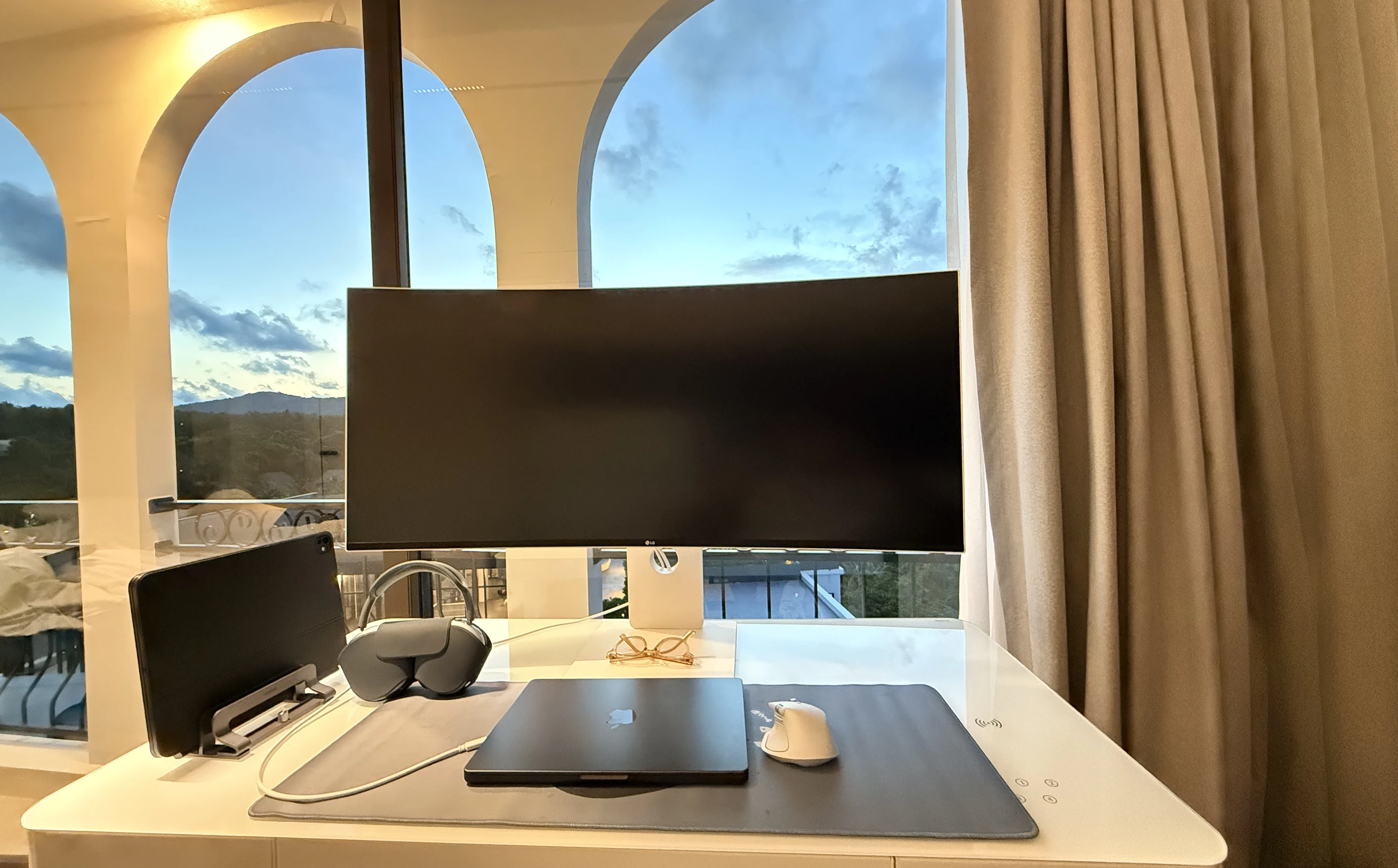 His desk at dusk: a curved monitor, a laptop, headphones and a tablet in a stand, in front of arched windows with hills beyond